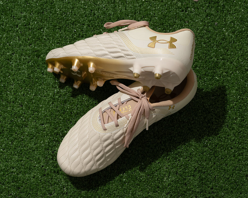 A pair of white/brown soccer cleats from Under Armour rest on brightly lit  artificial grass.
